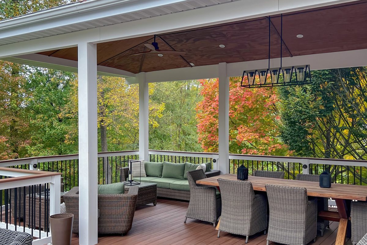 A luxury elevated deck with wicker patio furniture, wood dining table with chairs, ceiling fan and modern chandelier.  Pictured in autumn with fall colors.